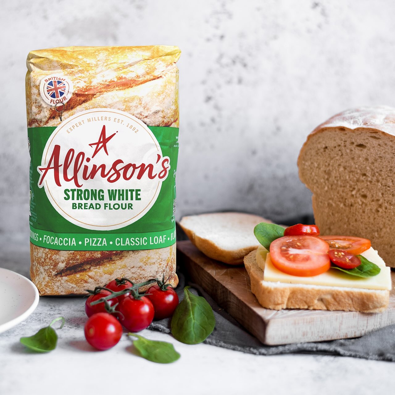 A freshly baked white loaf of bread cut into slices sits in the background. In the foreground, a cheddar and tomato sandwich is being made.